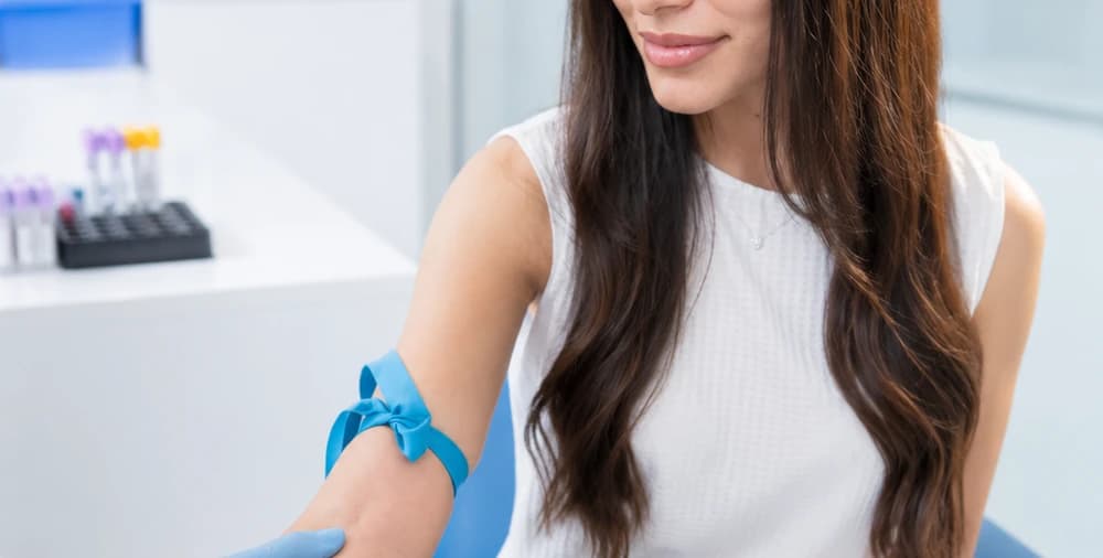 A seated person with an outstretched arm and a blue elastic band on the forearm, with a rack of sample tubes behind